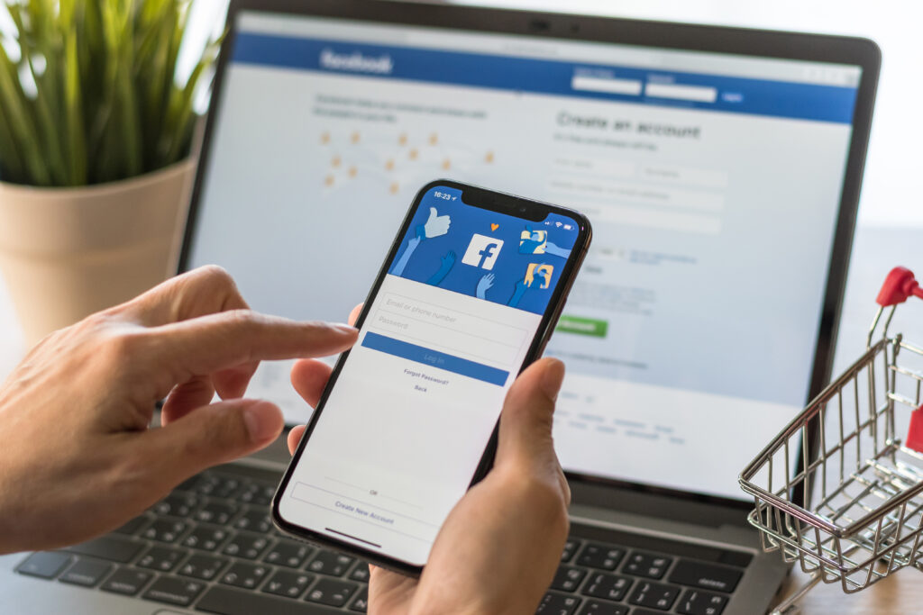 An image of a person holding a phone in front of a laptop. They are viewing a Facebook Ads post